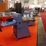 Industrielle Papierfaltmaschinen Stehen Auf Orangem Teppichboden Bei Einer Messe, Im Hintergrund Sind Menschen Zu Sehen.
