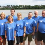 Sieben Erwachsene In Blauen T-Shirts Posieren Auf Einer Wiese Am Fluss Gemeinsam Für Ein Gruppenfoto.