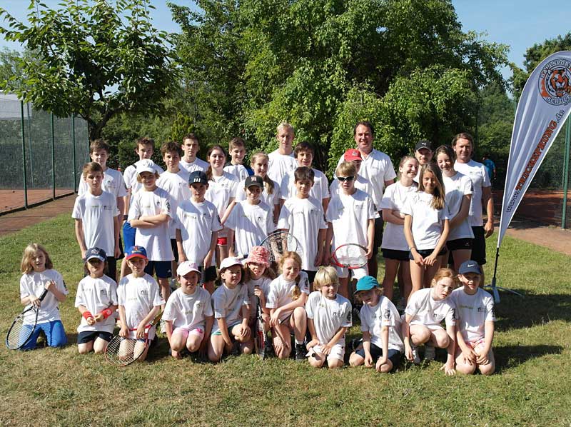 Kinder Und Erwachsene In Weißen Hemden Posieren Mit Tennisschlägern Nebeneinander Vor Tennisplätzen Im Freien.