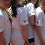 Vier Mädchen In Weißen &Quot;Hemmersbach Druck&Quot;-Shirts Stehen Zusammen Auf Einem Sonnigen Tennisplatz.
