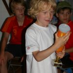Junge Im Weißen Hemd Hält Pokal, Zwei Weitere Jungen Sitzen Im Hintergrund Auf Einer Bank.