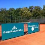 Tennissandplatz Mit Grünen Werbebannern Am Zaun, Dahinter Bäume Und Blauer Himmel.