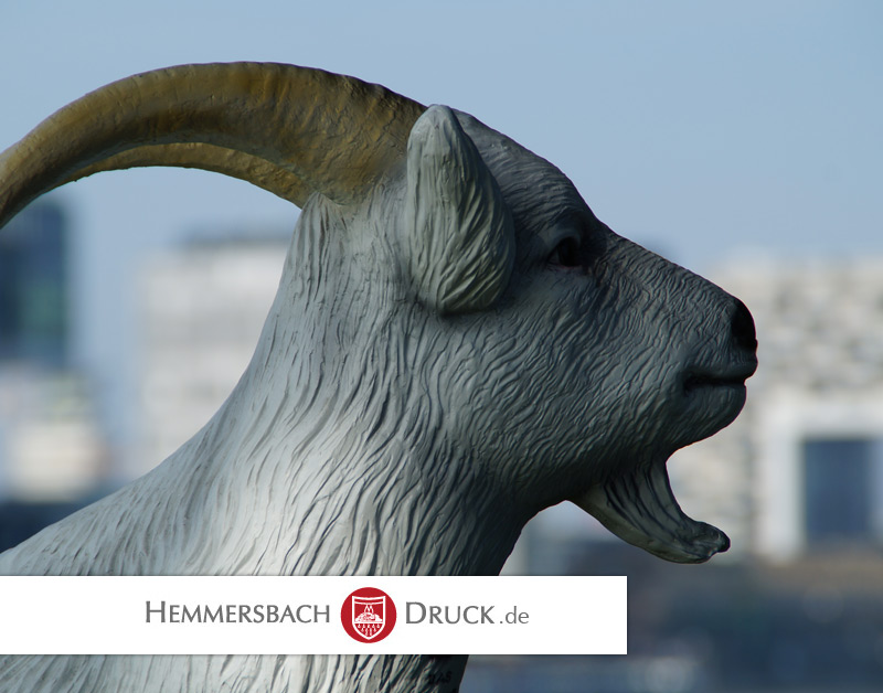 Ziegenskulptur Aus Metall Im Profil, Im Freien Vor Unscharfen Gebäuden, Unten Schriftzug &Quot;Hemmersbach Druck.de&Quot;.