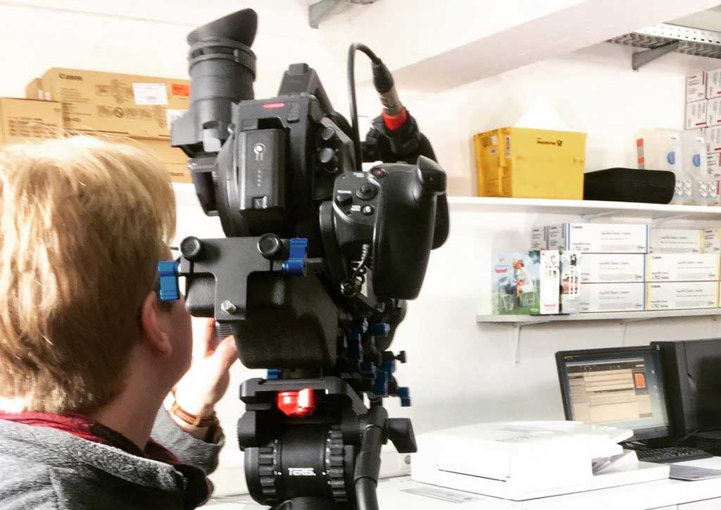 Person Filmt Mit Profi-Videokamera Einen Laptop Auf Einem Schreibtisch Im Büro Vor Regalen Mit Kartons.