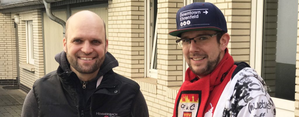 Zwei Männer Stehen Vor Einem Gebäude; Einer Trägt Schal Und Mütze, Der Andere Eine Schwarze Weste.
