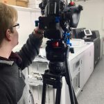 Person Bedient Professionelle Videokamera Auf Stativ In Raum Mit Großen Druck- Oder Kopiermaschinen.