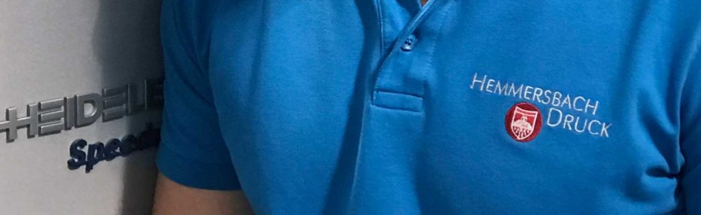 Person Im Blauen Hemmersbach Druck Polo Steht Neben Einer Heidelberg Speedmaster Druckmaschine In Einer Produktionshalle.