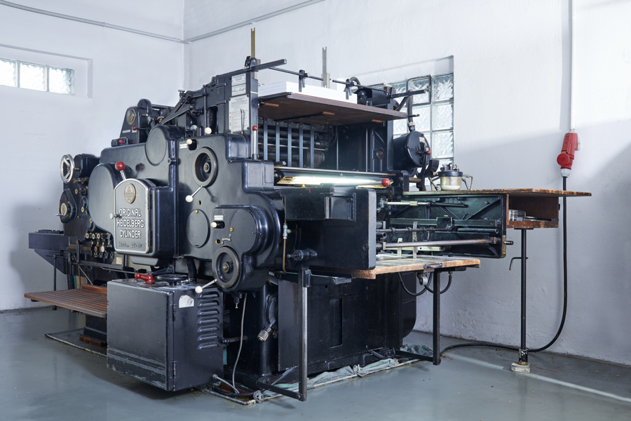 Große, Alte Industriebuchdruckmaschine Steht In Einem Raum Mit Weißen Wänden Und Betonboden.