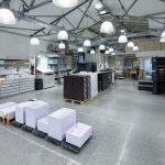 Druckmaschinen Stehen In Einer Modernen Druckerei Neben Gestapeltem Papier Und Regalen Unter Deckenbeleuchtung.