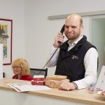 Mann Telefoniert Am Empfangstresen, Frau Sitzt Dahinter; Im Hintergrund Stehen Blumen.