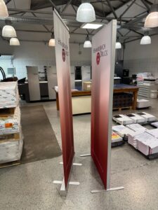 Zwei Hochformat-Banner Mit Aufdruck Stehen In Einer Druckerei Mit Papierstapeln Und Druckmaschinen.