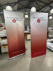 Zwei Rot-Weiße Banner Mit Logo Und Website Stehen In Einer Druckerei Neben Gestapeltem Papier.