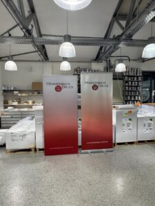 Zwei Werbebanner Stehen In Einer Druckerei Zwischen Papierstapeln Und Regalen Mit Arbeitsmaterialien.