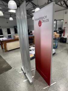 Zwei Roll-Ups Stehen In Einer Druckerei; Eines Mit Aufdruck &Quot;Hemmersbach Druck&Quot;, Das Andere Silbern Und Unbedruckt.