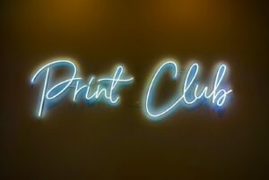 Leuchtreklame Mit Dem Schriftzug „Print Club“ In Schreibschrift Vor Einem Dunklen, Schlichten Hintergrund.