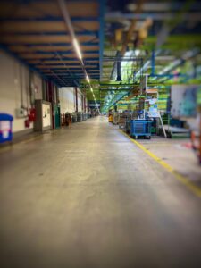 Ein Leerer, Breiter Gang In Einer Industriehalle Mit Regalen, Wagen Und Geräten An Den Seiten Unter Neonlicht.