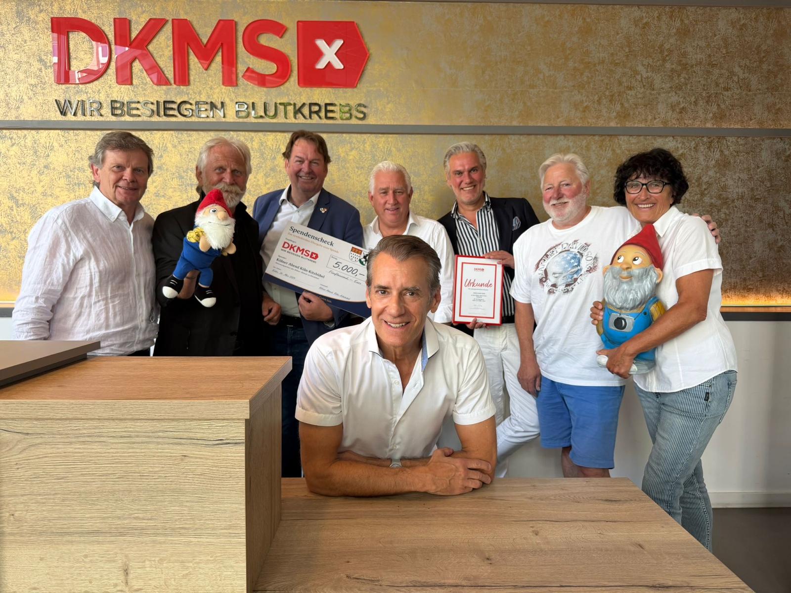 Acht Erwachsene Posieren Drinnen Vor Einem Dkms-Schild, Zwei Halten Gartenzwerge In Der Hand, Andere Halten Urkunden.