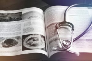 Drucksachen Für Arztpraxen Und Heilberufe In Köln: Terminkarten, Blöcke, Schilder Und Mehr Ein Stethoskop Liegt Auf Einem Aufgeschlagenen Medizinischen Lehrbuch Mit Bildern Und Text; Die Aufnahme Wurde Auf Einem Tisch Gemacht.
